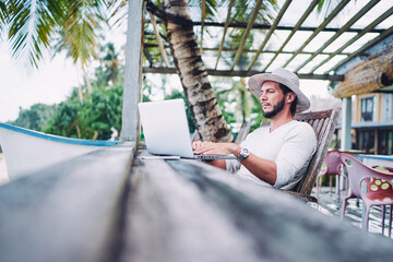 Technology and travel. Working outdoors. Freelance concept. Bearded young man using laptop in cafe on tropical beach.