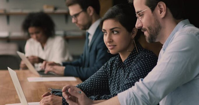 Indian Caucasian colleagues sit at shared coworking office brainstorm working together use laptop, talk, discuss business ideas cooperate use online application. Develop project, collaboration concept