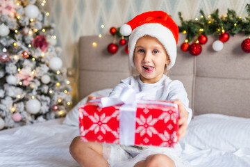 Preschool funny girl sit on bed near decorated Christmas tree in beautiful room in the holiday morning, enjoing with presents