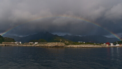 Naklejka na ściany i meble Beautiful multi-colored rainbow on the coast of Austvågøya island, Lofoten, Norway near village Svolvaer with traditional wooden drying flakes for stockfish and rugged mountains in background.