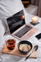 Breakfast in bed: a wooden tray with granola and cup of tea with a laptop on background.