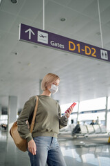 New normal and social distance concept. Young woman tourist wearing mask using smartphone searching airline flight status during corona virus 2019 outbreak at airport.
