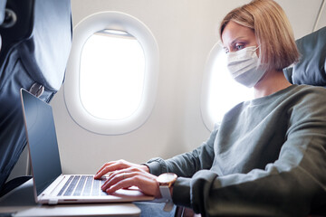 Young woman wearing face mask is traveling on airplane using laptop computer. New normal travel...