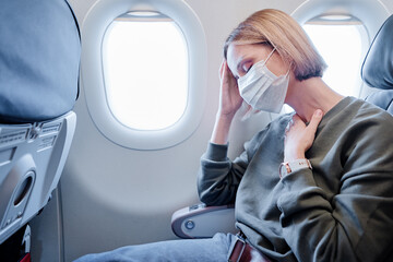 COVID-19 Young woman with face mask feeling unwell on plane. Fear of flying woman in airplane. Stress, headache, motion sickness and airsickness on plane during pandemic. - obrazy, fototapety, plakaty