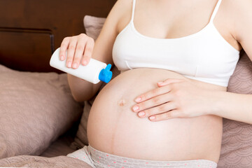 Fototapeta na wymiar a pregnant girl sits at home on the bed and smears smile an anti-stretch mark cream on her stomach. Pregnancy, motherhood, preparation and expectation concept