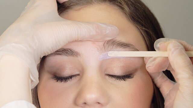 Correction of a shape of eyebrows with hot wax. Brow master applying wax on the eyebrow of female face. Wax correction of the shape of the eyebrows with spatula. Beauty industry. Close up. 