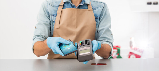 Cashier hand holding credit card reader machine and wearing disposable gloves, paying with smartphone during Covid-19 pandemic.