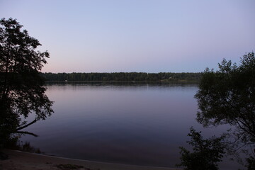 Beautiful view between the trees from riverbank on Volga river calm water, far shore with dark forest and clear blue sky at quiet summer night, Russian natural river landscape