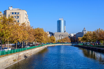 Fototapeta premium Landscape with Dambovita river, old buildings and yellow, orange and brown leaves in large trees in the center of Bucharest, Romania, in a sunny autumn day.