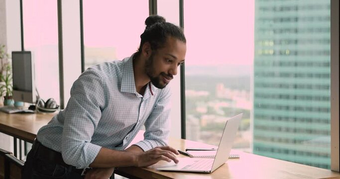 Serious focused african man using netbook in modern office room with city skyscrapers view. Mixed-race 35s start up owner of big prosperous enterprise working on laptop make research feels confident