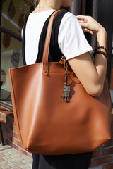 Cropped shot of a lady in white t-shirt, black jumpsuit and with brown leather bag on the shoulder. Retro keychain is fixed on the bag. The pendant is made as bronze robot on coiled leather cord. 