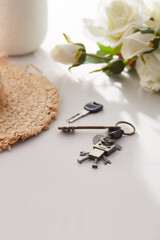 Subject shot of retro keychain made as bronze robot on coiled leather cord. The stylish keyring with charm and keys is located on the white table near straw hat and bunch of roses. 