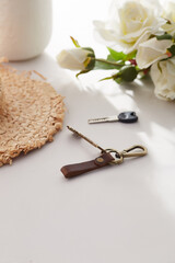 Subject shot of keychain with bronze lobster clasp, brown leather loop for belt and snap. The stylish keyring with keys is located on the white table near straw hat and bunch of roses. 