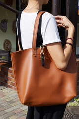 Cropped shot of a lady in white t-shirt, black jumpsuit and with brown leather bag on the shoulder. Bronze keyring with lobster clasp, black leather loop and snap is fixed on the bag. 