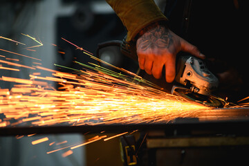 Cropped shot of tattooed metal worker using angle grinder while polishing metallic profile with sparks 
