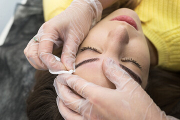 In a beauty salon, a master cosmetologist makes permanent makeup for a European girl, wipes her eyebrows with a cotton pad.