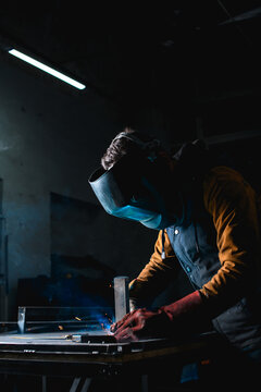 Welder in mask and gloves working with metal profile in unlit workshop 