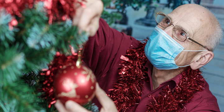Male aduld person in eyewear and medical face mask decorate Christmas tree with red toy.