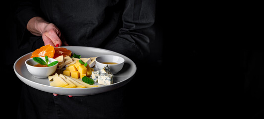 chef in a black suit holds in his hands Cheese plate on Dark grey black background