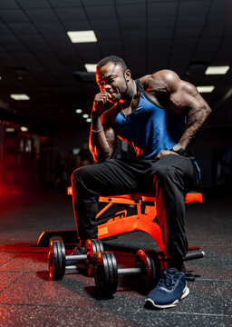Handsome african bodybuider with perfect body. Male sportsman sits on bech with dumbbell near legs.