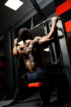 Athlete pumps biceps on the simulator. African handsome bodybuilder in gym. View from the back.