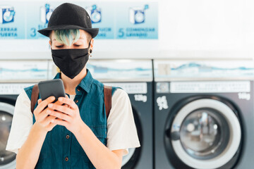 Bored young woman chatting with her friends via her smartphone while waiting for her laundry to be...
