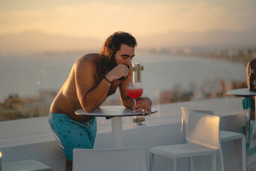 Bearded man with swim shorts and no shirt is drinking a red colored cocktail at the skybar of a hotel in s'Arenal, Mallorca during sunset