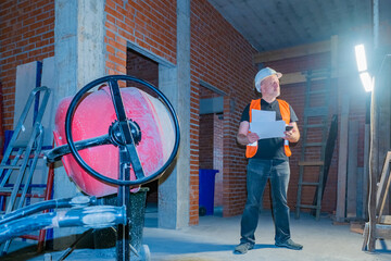 A man in an orange vest in a building under construction. Engineer with drawings and phone in his hands.The foreman inspects a suspended searchlight in a house under construction. Construction concept