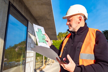 The engineer examines the drawings. A man with papers and a phone near a house under construction. Construction of country houses. The customer at the construction site.