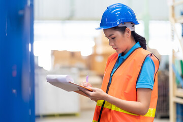 Asian female warehouse worker with safety vest and helmet working and using clipboard for checking products or parcel goods on shelf pallet in storage warehouse.  people, warehouse, industry concept