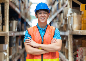 Confident smiling Asian male warehouse worker in safety vest and helmet standing with arms crossed...