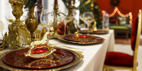 Naklejka premium Beautiful served table with decorations, candles and lanterns. The living room is decorated with lights and a Christmas tree. Holiday setting. decorative golden horse on a plate.
