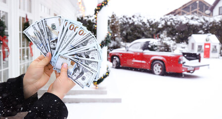 female hand holds a fan of dollars. against the background of a red car and a house. money for christmas. win money for christmas and new year