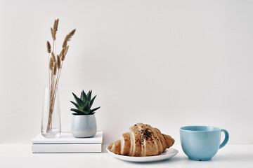 Croissant and a mug, a vase and a pot with a plant on the books, white background. Eco-friendly materials in interior decor, minimalism. Copy space, mock up.