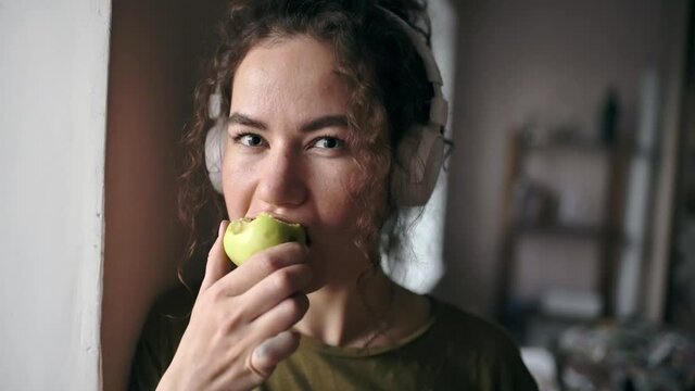 Young happy girl in headphones eats green apple and looks into the frame