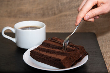 A woman's hand holds a dessert metal spoon and takes a piece of chocolate cake next to a Cup of coffee. The concept of sweet coffee break with cake
