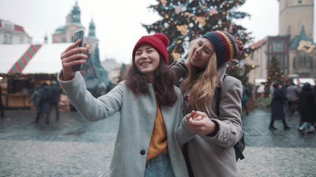 Portrait of two girls making selfie in center of square in european town. Girls taking photo in heart of christmas fare. Background of christmas tree and decorations. Concept of travel, gadgets.