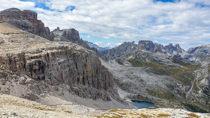 A panoramic view on Dolomites in Italy. There are sharp and steep mountain slopes around. At the bottom of a small valley there is a small navy blue lake. The sky is full of soft clouds. Raw landscape