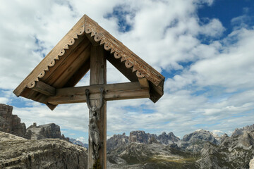 A wooden cross with a figure of Jesus on it with panoramic view on Dolomites in Italy. There are sharp and steep mountain slopes around. The sky is full of soft clouds. Spirituality and meditation