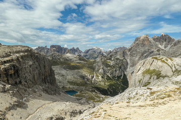 A panoramic view on Dolomites in Italy. There are sharp and steep mountain slopes around. At the bottom of a small valley there is a small navy blue lake. The sky is full of soft clouds. Raw landscape
