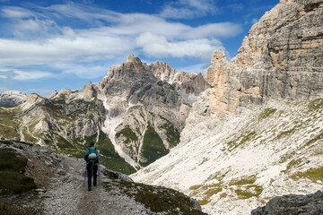 Fototapeta premium A woman with backpack and sticks hiking on a path in Italian Dolomites. There are sharp and steep mountains around. Lower slopes of the mountains overgrown with small, green plants. Raw landscape