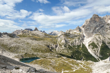 A panoramic view on Dolomites in Italy. There are sharp and steep mountain slopes around. At the bottom of a small valley there is a small navy blue lake. The sky is full of soft clouds. Raw landscape
