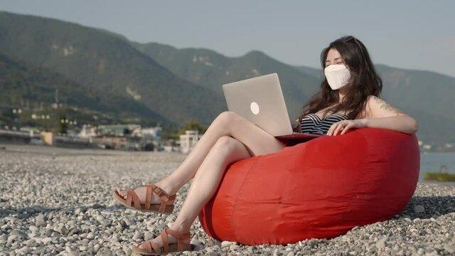 Young business woman in bikini in medical mask working on laptop alone by the sea at the beach. Freelance or distant working during pandemic. Coronavirus protection and social distancing concept.