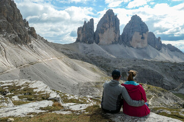 Naklejka premium A couple in hiking outfit, sitting on the ground and enjoying the view on the famous Tre Cime di Lavaredo (Drei Zinnen) in Italian Dolomites. Desolated and raw landscape, full of lose stones. Overcast