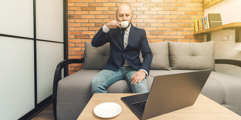 Attractive bearded bald man sitting on sofa and drinking coffee, looking at laptop, modern interior loft design