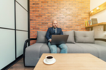 Bearded bald man, businessman or freelancer sitting on sofa and working on laptop from home, modern interior loft design