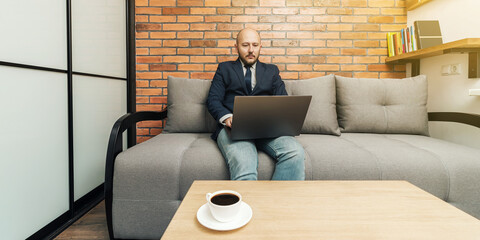 Bearded bald man, businessman or freelancer sitting on sofa and working on laptop from home, modern interior loft design