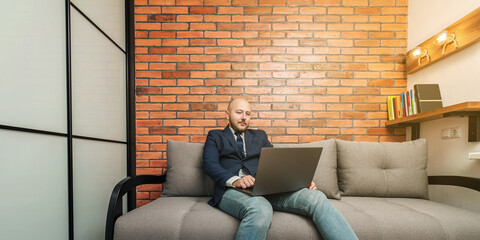 Bearded bald man, businessman or freelancer sitting on sofa and working on laptop from home, modern interior loft design