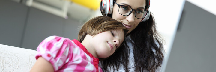 Beautiful woman in glasses and headphones work for laptop. Daughter in plaid shirt put her head on mother shoulder and look at screen.