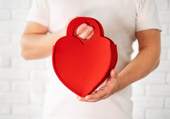 Caucasian man in a white t-shirt, holding a purse gift in the form of a red heart. Men's hands close-up. A gift for Valentine's day. Valentine card